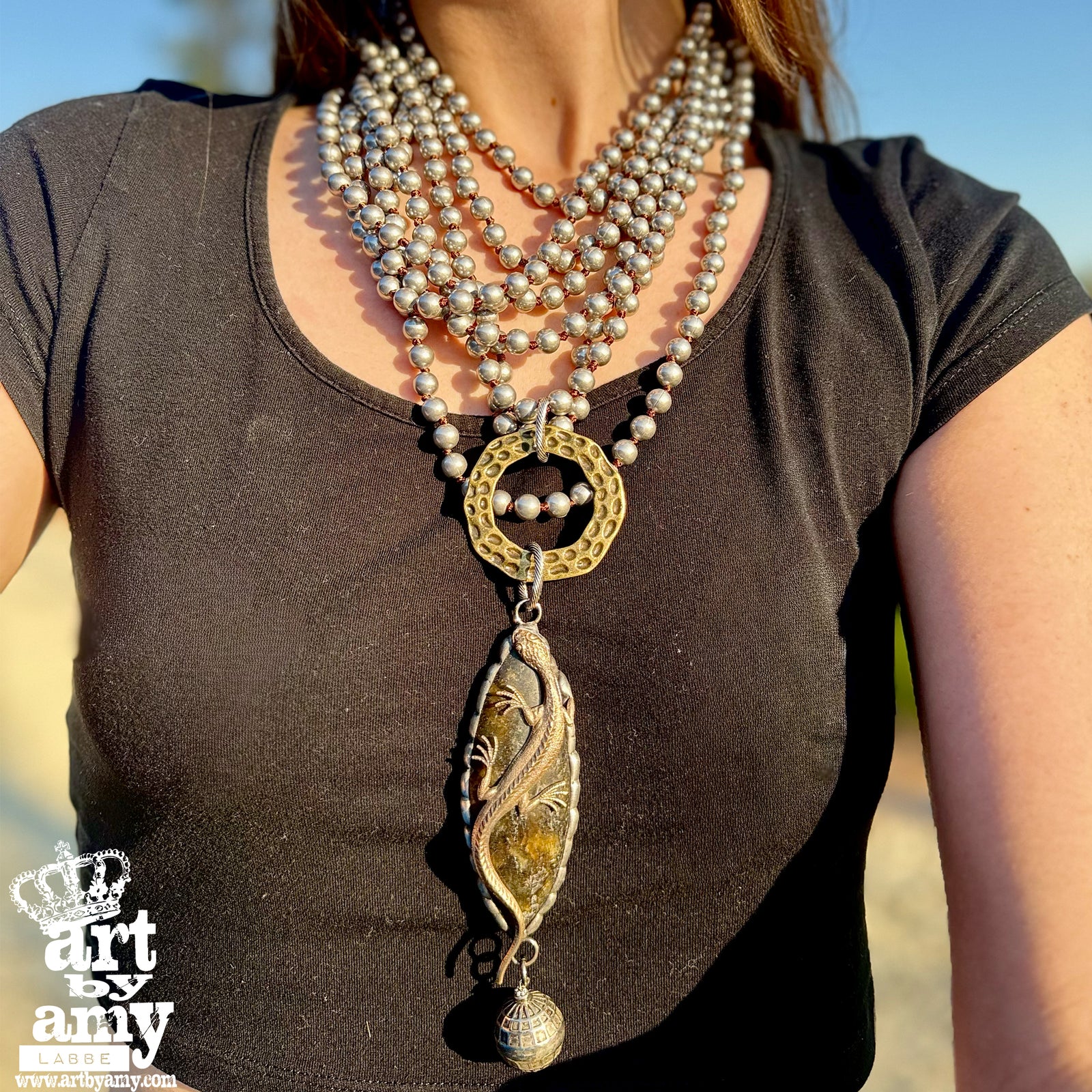 Person wearing a long, ornate necklace with a large pendant against a blurred natural background.