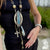 Person wearing a long, ornate necklace with a blue stone pendant outdoors.